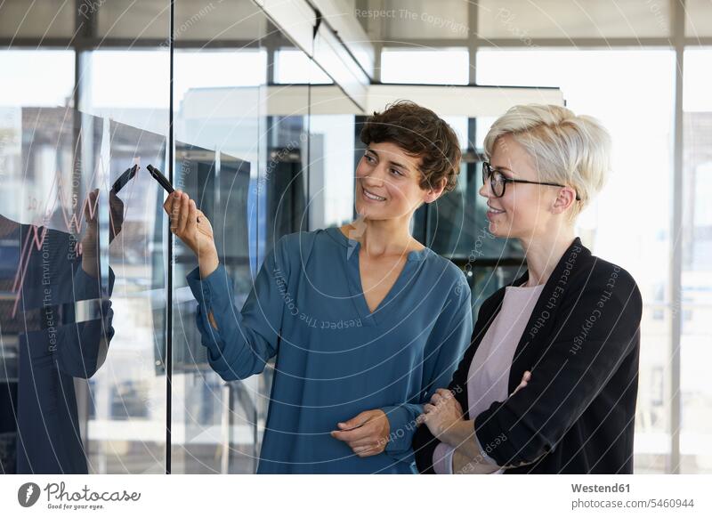 Zwei lächelnde Geschäftsfrauen mit Blick auf Diagramm auf Glasscheibe im Büro Fenster Grafik Graph Grafiken Chart ansehen Office Büros Businesswomen