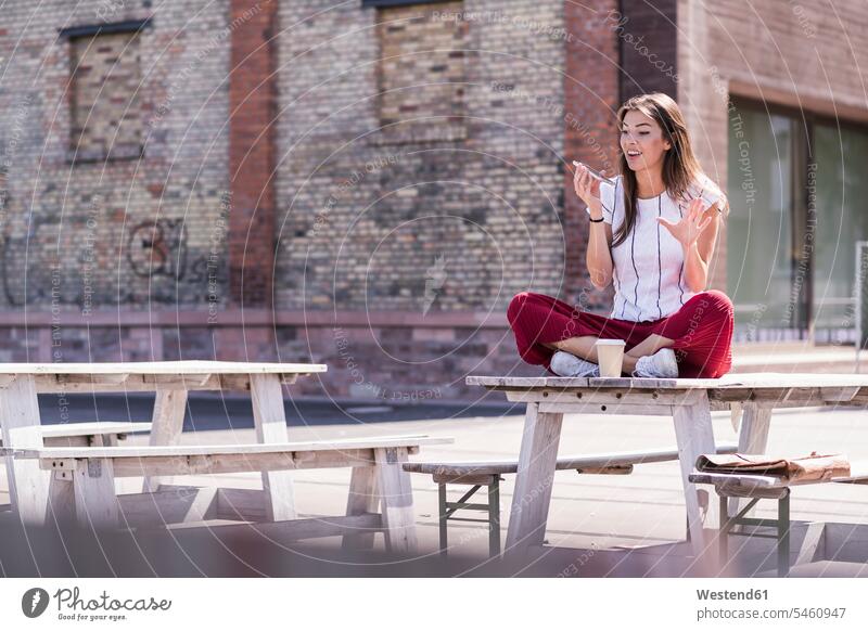Junge Frau sitzt auf einem Tisch in einem Biergarten und benutzt ein Mobiltelefon Leute Menschen People Person Personen Europäisch Kaukasier kaukasisch 1 Ein