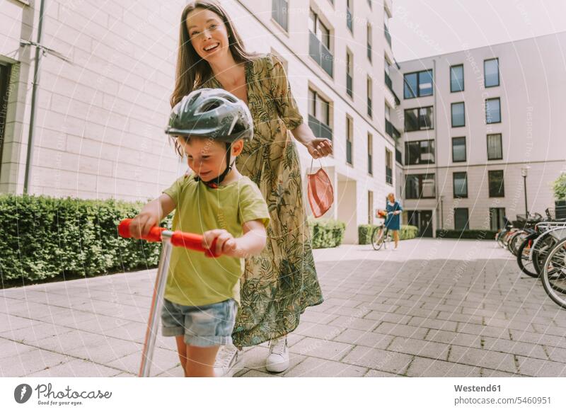 Lächelnde Mutter mit Gesichtsmaske und Sohn mit Roller in der Stadt Kinderfahrzeuge Tretroller Miteinander Zusammen Gesund außen draußen im Freien staedtisch