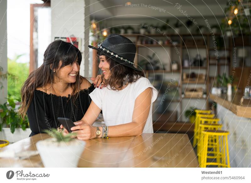 Zwei Freundinnen lächeln sich im Café mit Smartphone an Frau weiblich Frauen Cafe Kaffeehaus Bistro Cafes Cafés Kaffeehäuser iPhone Smartphones Freunde