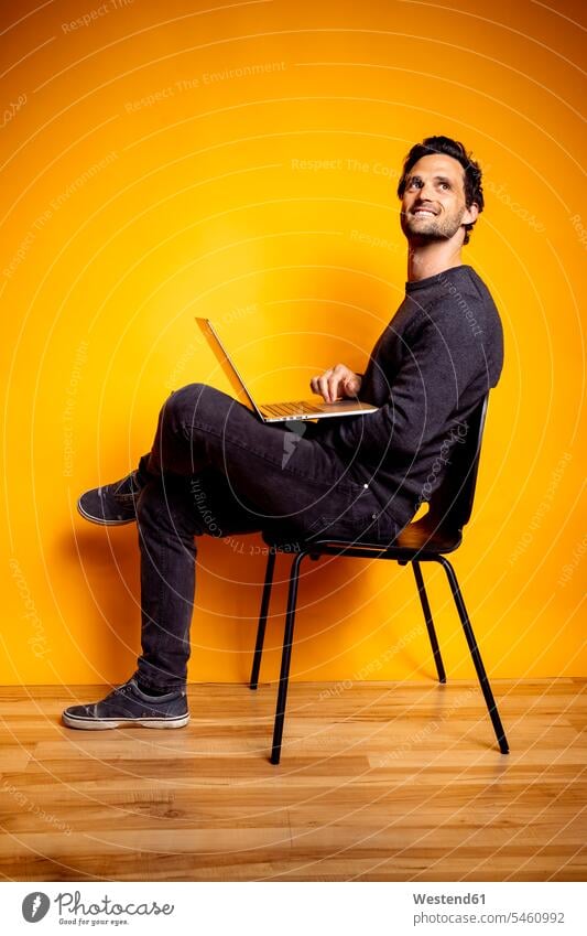 Hübscher Mann schaut weg, während er mit Laptop auf einem Stuhl sitzt Farbaufnahme Farbe Farbfoto Farbphoto farbiger Hintergrund Studioaufnahme Studioaufnahmen