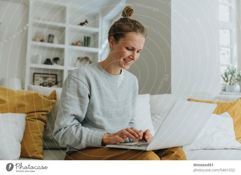 Lächelnde Frau benutzt Laptop, während sie zu Hause auf dem Sofa sitzt Farbaufnahme Farbe Farbfoto Farbphoto Innenaufnahme Innenaufnahmen innen drinnen Tag