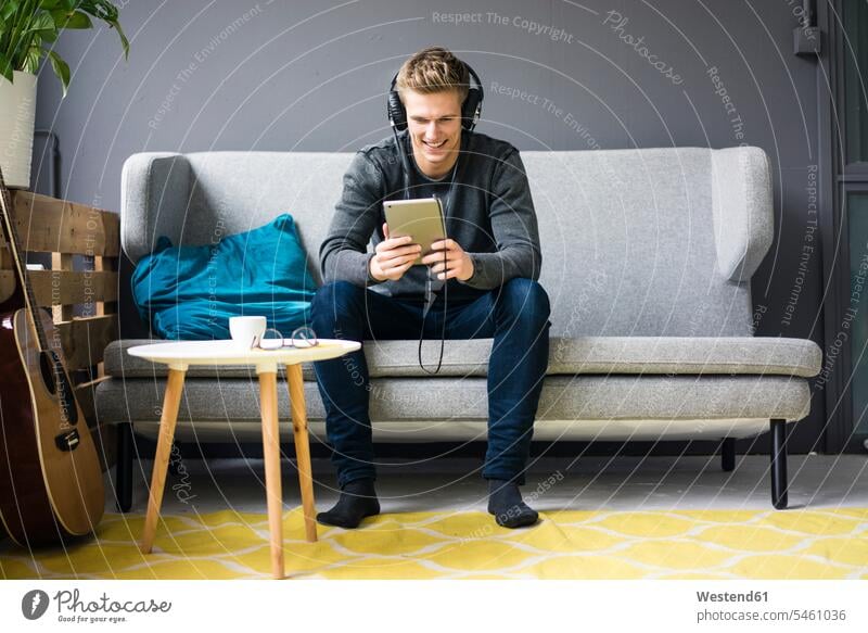 Lächelnder junger Mann mit Gitarre, Tablet und Kopfhörer sitzt auf der Couch Gitarren sitzen sitzend lächeln Männer männlich Tablet Computer Tablet-PC Tablet PC