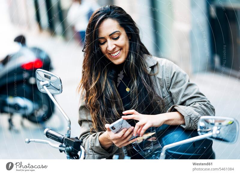 Lächelnd Biker Frau mit Handy auf Motorrad in der Stadt Farbaufnahme Farbe Farbfoto Farbphoto Außenaufnahme außen draußen im Freien Tag Tageslichtaufnahme