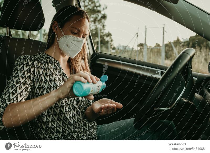 Mittlere erwachsene Frau mit Schutzmaske, die im Auto Desinfektionsmittel verwendet Flaschen KFZ Verkehrsmittel Automobil Autos PKW PKWs Wagen sitzend sitzt