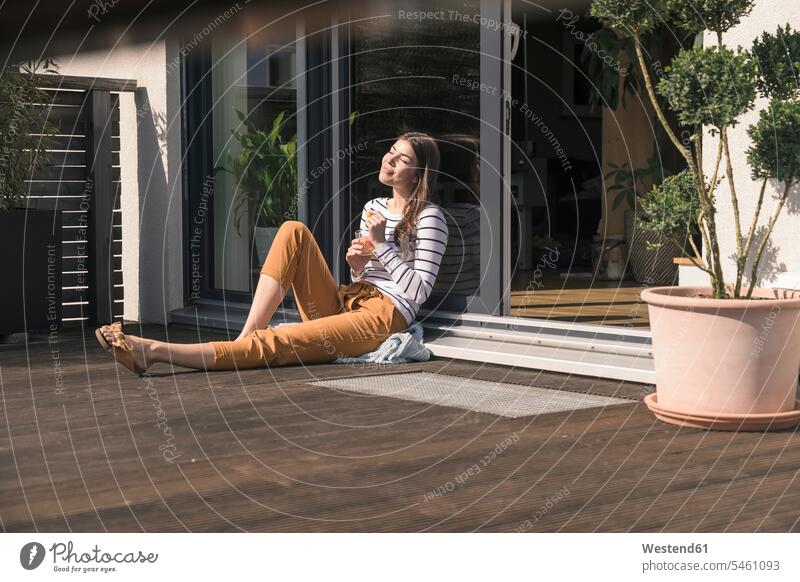 Junge Frau sitzt auf der Terrasse zu Hause und genießt den Sonnenschein Deutschland auf dem Boden sitzen auf dem Boden sitzend bequem Streifen gestreift