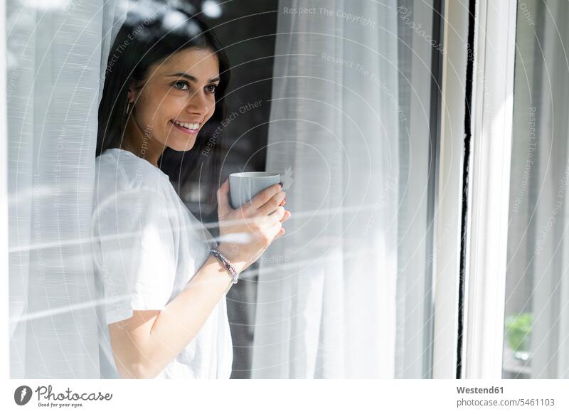Lächelnde junge Frau mit Tasse Kaffee hinter Fensterscheibe Leute Menschen People Person Personen Alleinstehende Alleinstehender Singles Unverheiratete