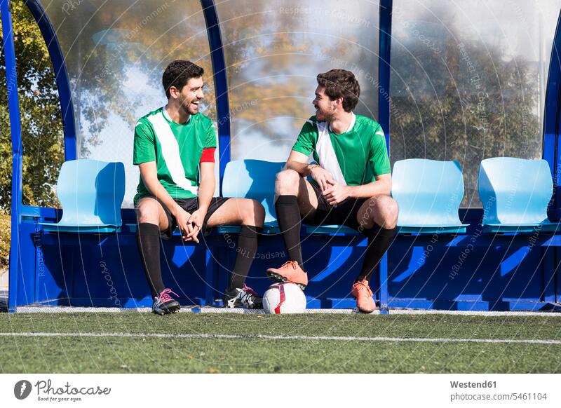 Zwei glückliche Fußballspieler sitzen auf einer Bank am Fußballplatz und unterhalten sich Glück glücklich sein glücklichsein Fussballspieler Fussballer