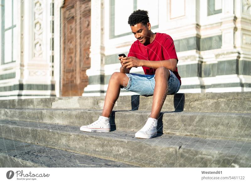 Junger Mann sitzt auf einer Außentreppe und überprüft sein Smartphone, Florenz, Italien Leute Menschen People Person Personen Afrikanisch