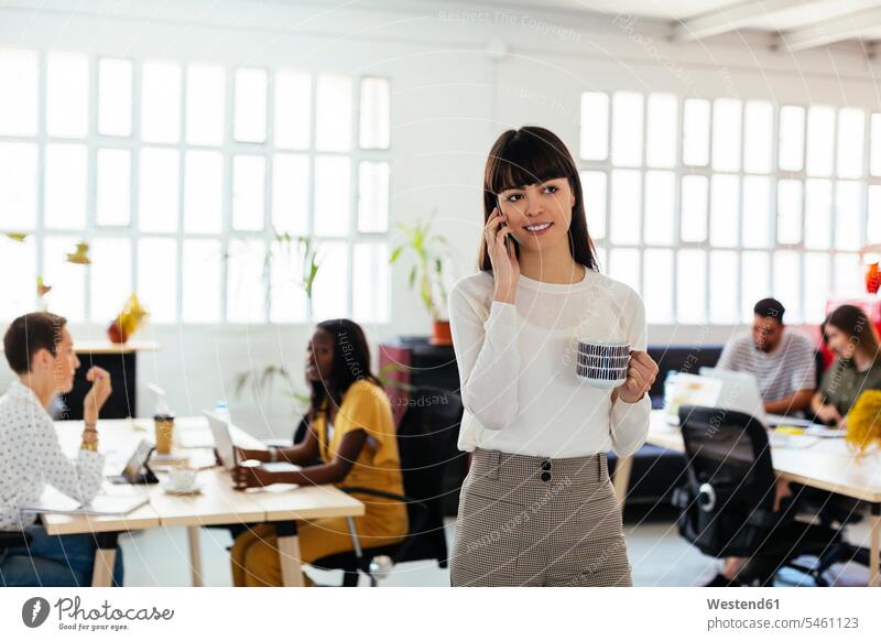 Porträt einer lächelnden jungen Frau am Handy im Büro mit Kollegen im Hintergrund Portrait Porträts Portraits Arbeitskollegen telefonieren anrufen Anruf