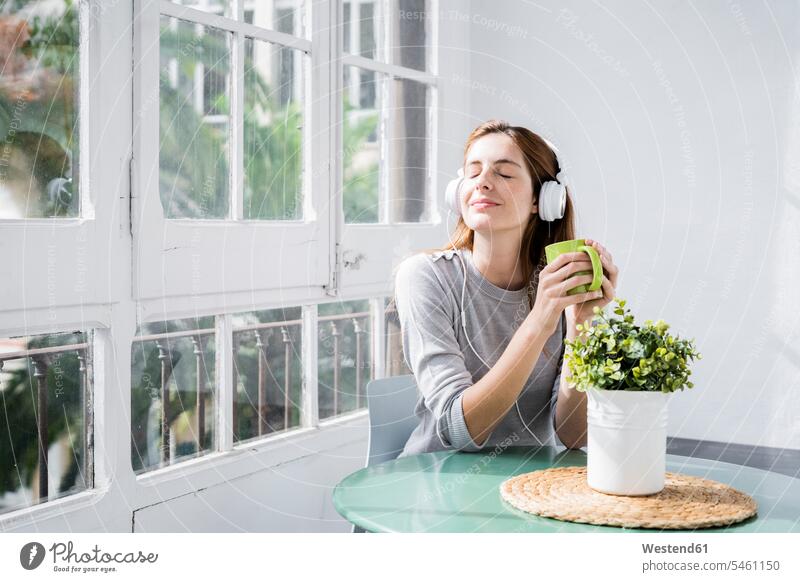 Junge Frau stting am Fenster zu Hause mit Kaffeetasse und Kopfhörer Leute Menschen People Person Personen Alleinstehende Alleinstehender Singles Unverheiratete