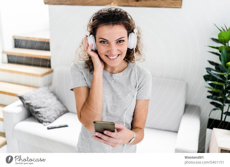 Porträt einer lächelnden jungen Frau, die mit Kopfhörer und Handy Musik hört T-Shirts Couch Couches Liege Sofas Kopfhoerer Telekommunikation Handies Handys