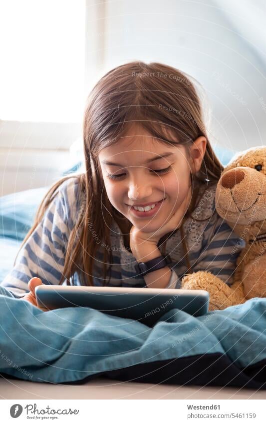 Porträt eines lächelnden Mädchens mit Teddybär auf dem Bett liegend mit digitalem Tablett Betten Teddies Teddybaer Teddybaeren Teddybären Medium entspannen