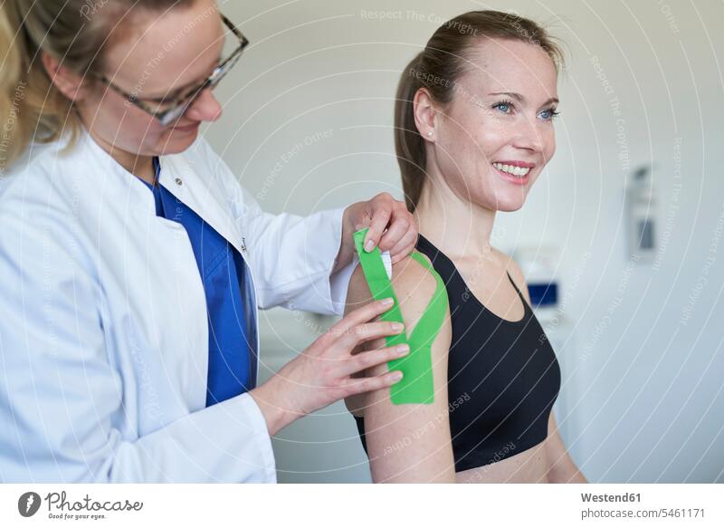 Ärztin beim Anlegen von Klebeband an der Schulter eines Patienten in einer Arztpraxis Deutschland Arztbesuch Arztbesuche Arztkittel Vorbeugung Sachverstand