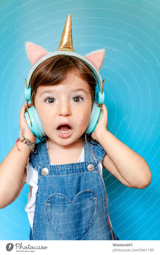 Porträt eines süßen kleinen Mädchens, das mit einhornförmigen Kopfhörern auf blauem Hintergrund Musik hört und singt Leute Menschen People Person Personen