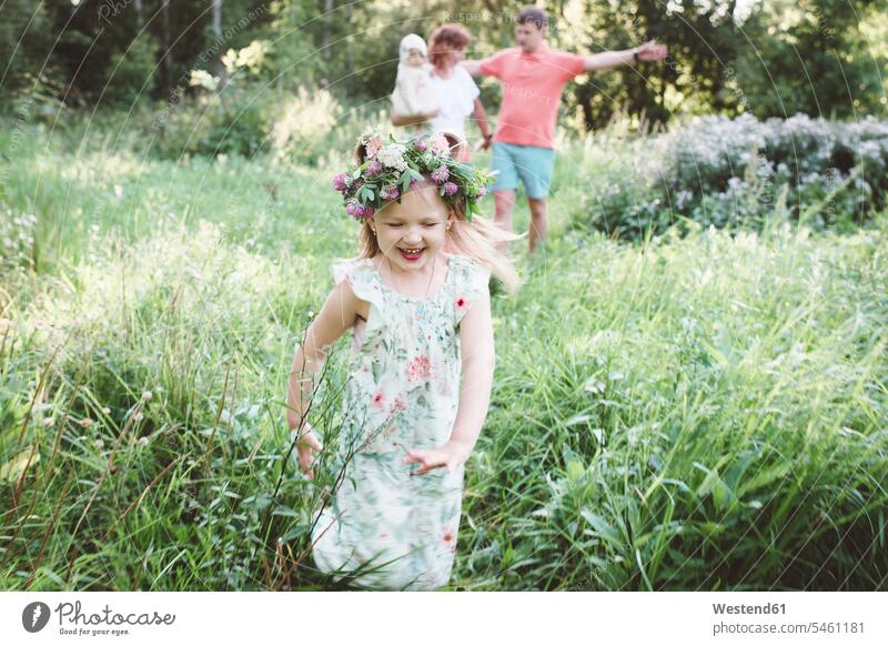 Glückliches kleines Mädchen mit Blumenkranz, das in der Natur vor seinen Eltern wegläuft Leute Menschen People Person Personen Europäisch Kaukasier kaukasisch