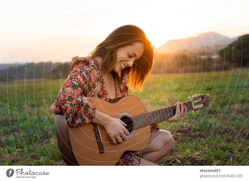 Lächelnde junge Frau spielt Gitarre auf der Wiese bei Sonnenuntergang Farbaufnahme Farbe Farbfoto Farbphoto Außenaufnahme außen draußen im Freien