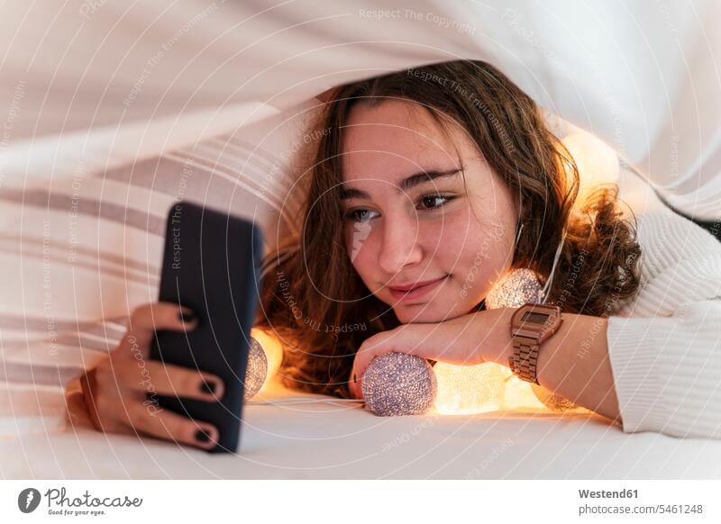 Lächelndes Teenager-Mädchen mit Lichterkette, das sein Handy unter der Bettdecke benutzt Betten Telekommunikation telefonieren Handies Handys Mobiltelefon