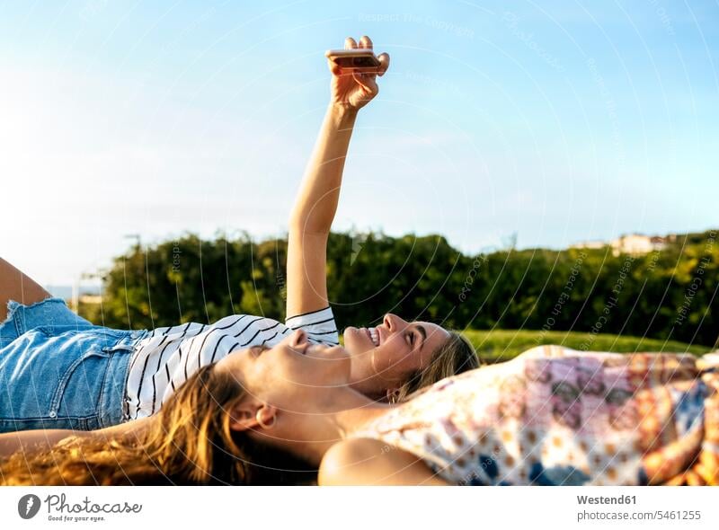 Lächelnde junge Frau, die ein Selfie mit einer Freundin macht, während sie sich auf einem Feld an einem sonnigen Tag entspannt Farbaufnahme Farbe Farbfoto
