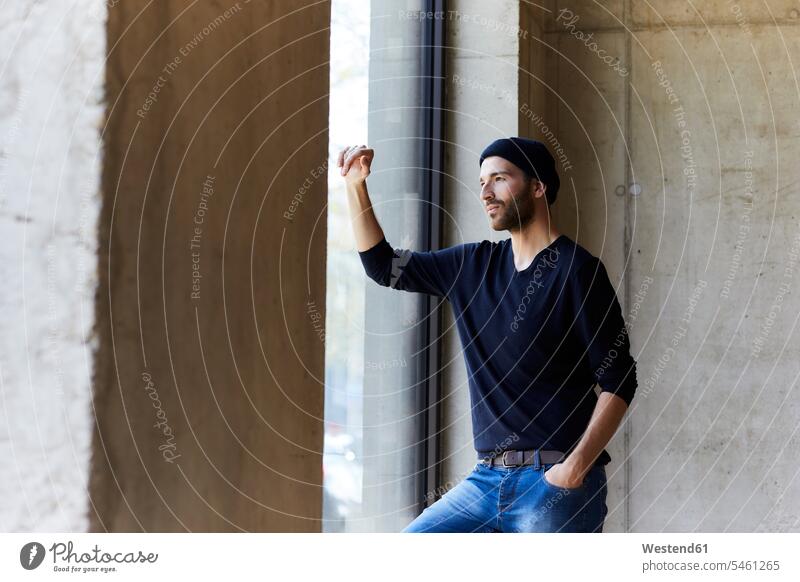 Junger Mann mit Mütze schaut aus dem Fenster Deutschland Betonwand Betonwände Betonwaende denken nachdenken überlegen Verträumt träumerisch Lifestyle Lebensstil