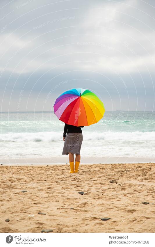 Frau mit bunten Regenschirm sitzt am Strand Regenbogenfarben Spektralfarben Beach Straende Strände Beaches weiblich Frauen multikulturell Toleranz tolerant