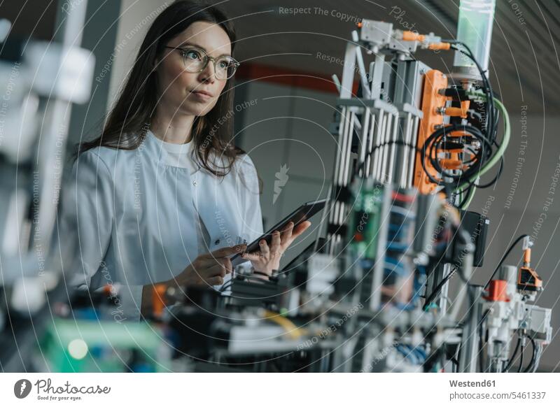 Wissenschaftlerin hält ein digitales Tablett in der Hand, während sie Maschinen im Labor untersucht Farbaufnahme Farbe Farbfoto Farbphoto Innenaufnahme