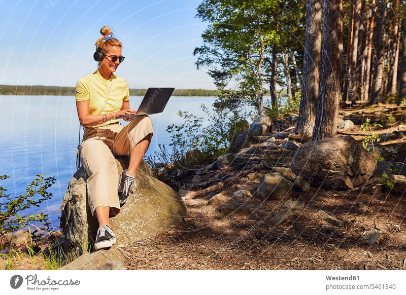 Mittelgroße erwachsene Frau mit Sonnenbrille und Laptop am See im Tiveden-Nationalpark, Schweden Farbaufnahme Farbe Farbfoto Farbphoto Skandinavien