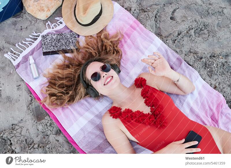 Glückliche junge Frau liegt auf einem Handtuch am Strand und hört Musik Leute Menschen People Person Personen gelockt gelockte Haare gelocktes Haar lockig