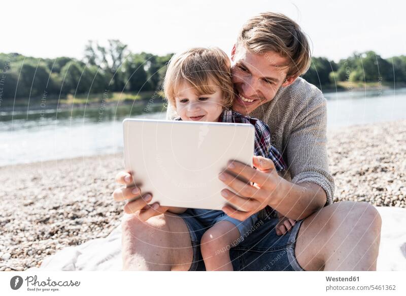 Vater und Sohn spielen am digitalen Tablet am Flussufer Söhne Tablet Computer Tablet-PC Tablet PC iPad Tablet-Computer Papas Väter Vati Vatis Papis Sommer
