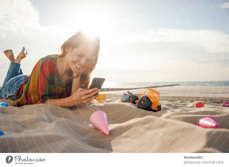 Rothaarige Frau liegt mit Strandspielzeug am Strand und benutzt ein Smartphone Beach Straende Strände Beaches iPhone Smartphones weiblich Frauen spielen