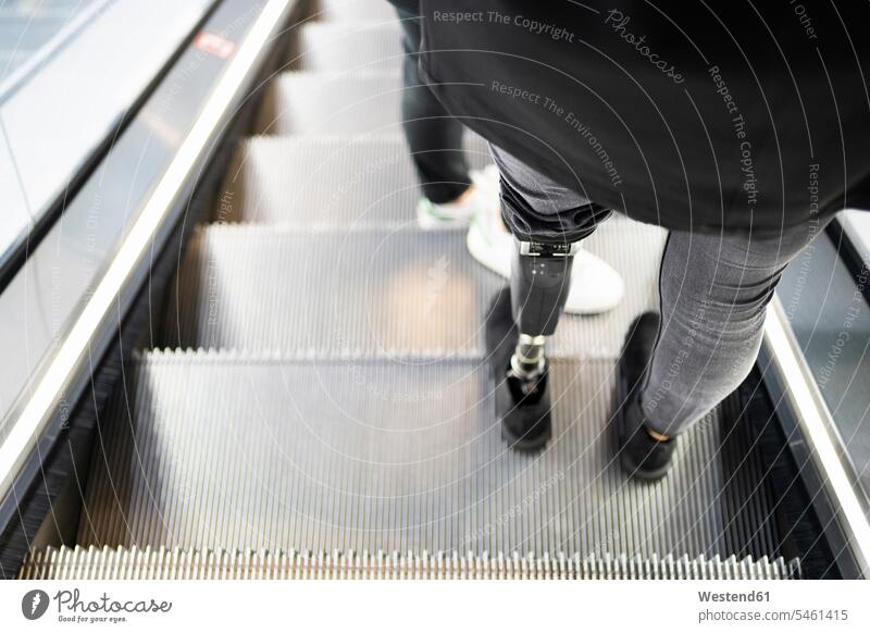 Nahaufnahme einer auf der Rolltreppe stehenden Frau mit Beinprothese Fahrtreppe steht Miteinander Zusammen mobil auf Achse in Bewegung Rueckansicht