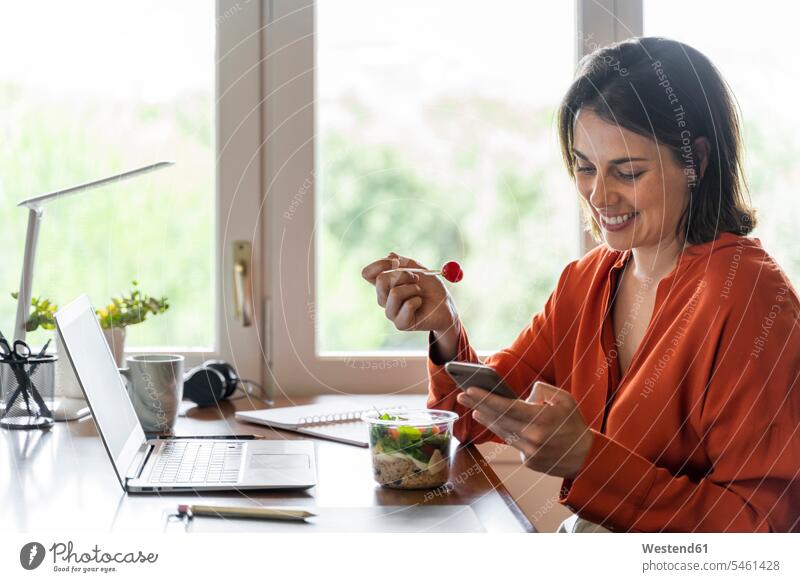Lächelnde Geschäftsperson, die ein Mobiltelefon benutzt, während sie zu Hause Salat isst Farbaufnahme Farbe Farbfoto Farbphoto Innenaufnahme Innenaufnahmen