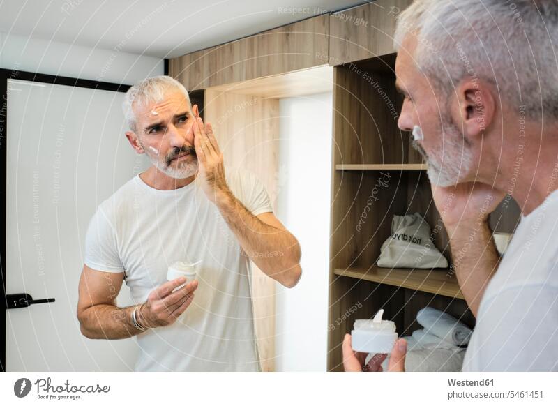 Reifer Mann, der Gesichtscreme aufträgt, während er in den Spiegel im Badezimmer schaut Farbaufnahme Farbe Farbfoto Farbphoto Innenaufnahme Innenaufnahmen innen