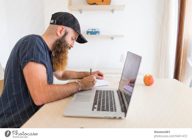 Bärtiger junger Mann sitzt am Schreibtisch mit Laptop und schreibt etwas auf Arbeitstisch Schreibtische schreiben aufschreiben notieren schreibend Schrift Bart