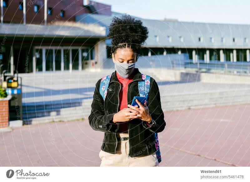 Student trägt eine schützende Gesichtsmaske und benutzt ein Smartphone, während er auf der Straße in der Stadt steht Farbaufnahme Farbe Farbfoto Farbphoto