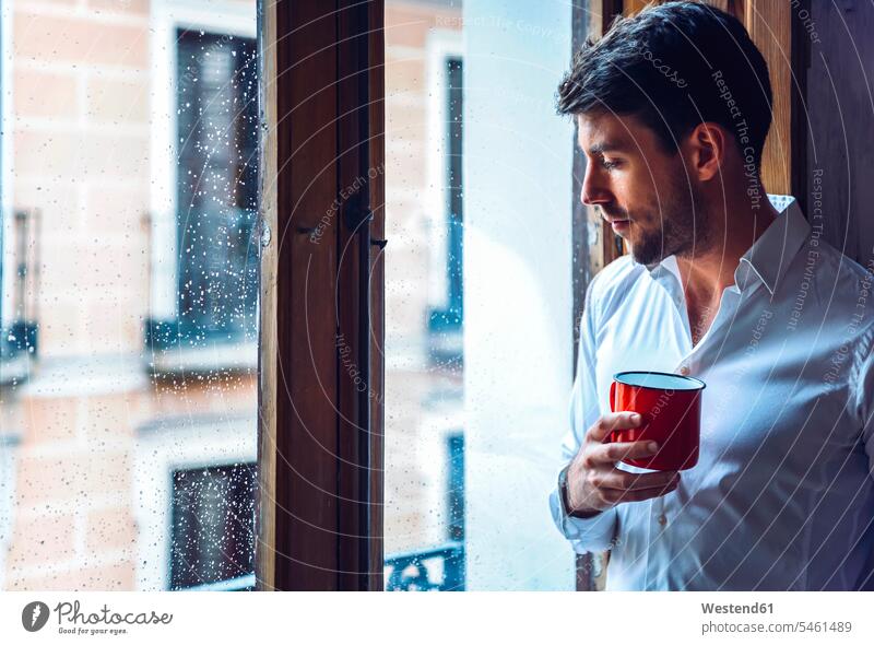 Junger Mann hält Tasse und schaut aus dem Fenster Leute Menschen People Person Personen Alleinstehende Alleinstehender Singles Unverheiratete Unverheirateter