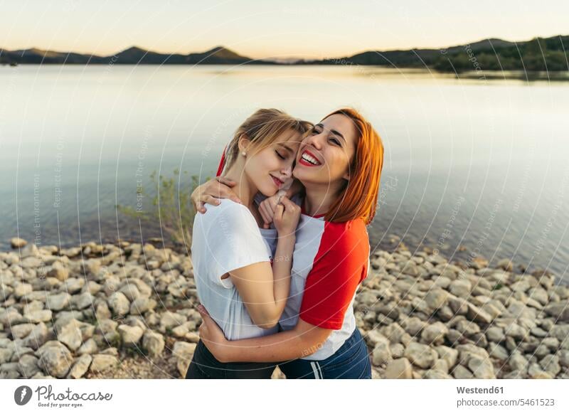 Glückliche Frau umarmt Freundin während des Sonnenuntergangs am See stehend Farbaufnahme Farbe Farbfoto Farbphoto Außenaufnahme außen draußen im Freien