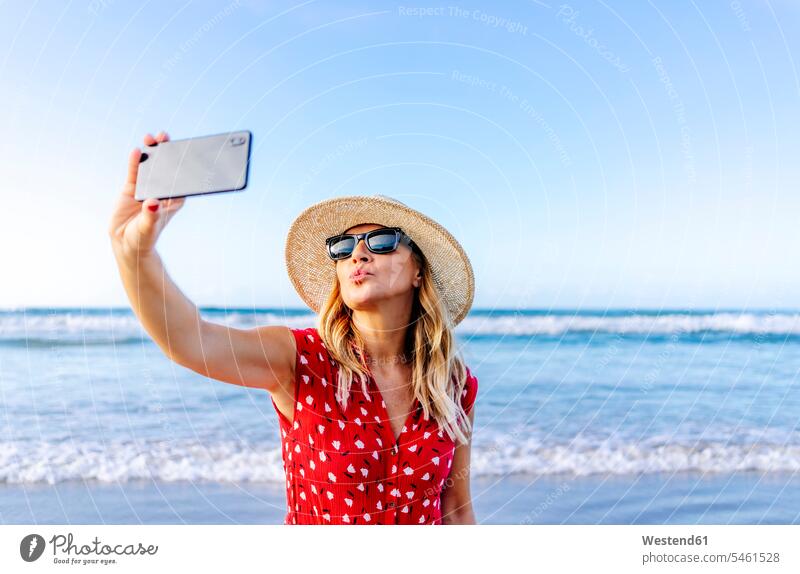 Blonde Frau in rotem Kleid und Hut, die ein Smartphone benutzt und ein Selfie am Strand macht, wobei sie einen Kussmund macht Telekommunikation telefonieren