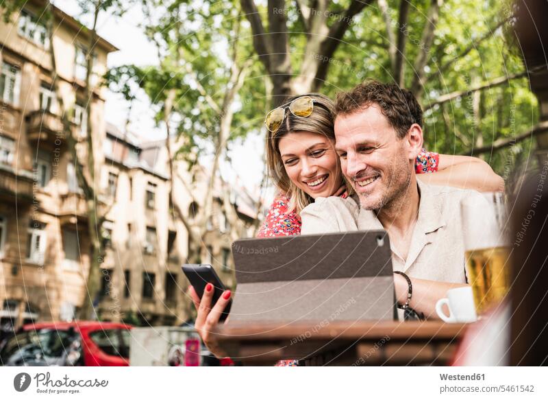Lächelndes Paar, das sich Tablet und Handy in einem Café im Freien teilt Cafe Kaffeehaus Bistro Cafes Cafés Kaffeehäuser Teilen Sharing lächeln glücklich Glück