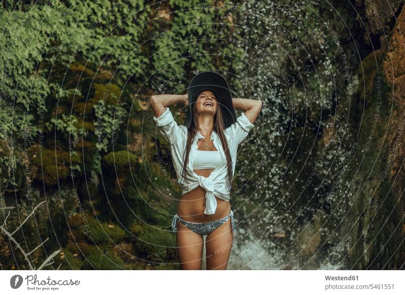 Glückliche junge Frau mit Hut, die in der Nähe eines Wasserfalls steht Badebekleidung Bikinis Hüte sommerlich Sommerzeit entspannen relaxen entspanntheit relaxt