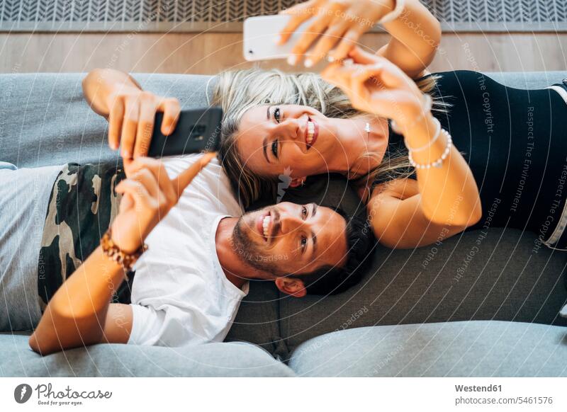 Porträt eines lachenden Paares, das auf der Couch liegt und Selfies mit Smartphones macht Leute Menschen People Person Personen Europäisch Kaukasier kaukasisch