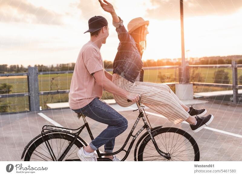 Glückliches junges Paar zusammen auf einem Fahrrad bei Sonnenuntergang auf dem Parkdeck Hüte Raeder Räder Bike Bikes Fahrräder Rad sitzend sitzt abends