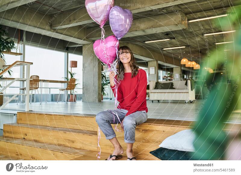Geschäftsfrau hält Luftballons in der Hand, während sie im Loft-Büro auf einer Treppe sitzt Farbaufnahme Farbe Farbfoto Farbphoto Innenaufnahme Innenaufnahmen