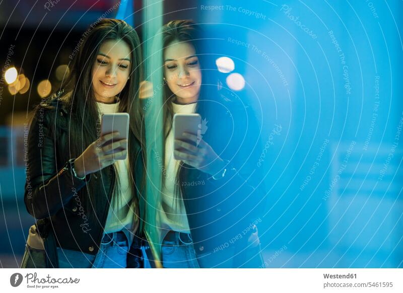 Porträt eines lächelnden Teenager-Mädchens, das sich an eine Glasscheibe lehnt und auf ein Smartphone schaut Leute Menschen People Person Personen Europäisch