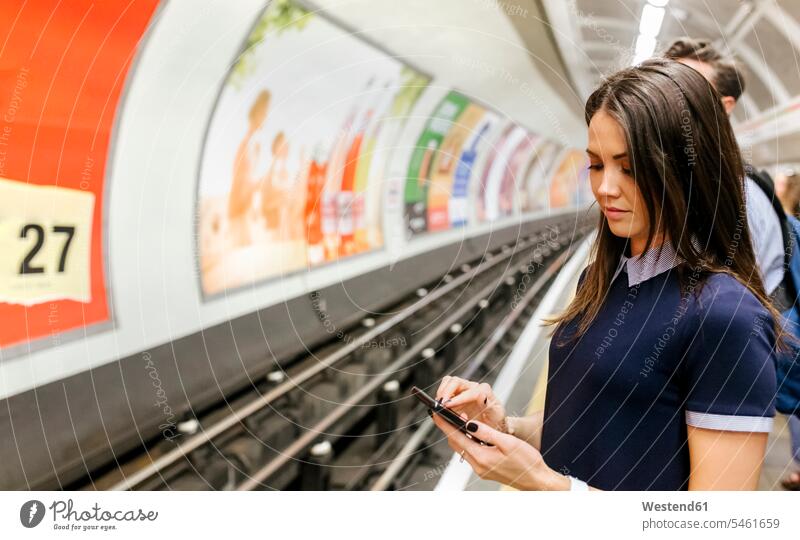 UK, London, junge Frau wartet auf U-Bahn-Station Plattform Blick auf Handy Smartphone iPhone Smartphones schauen sehend warten U-Bahnhofsbahnsteig Bahnsteig
