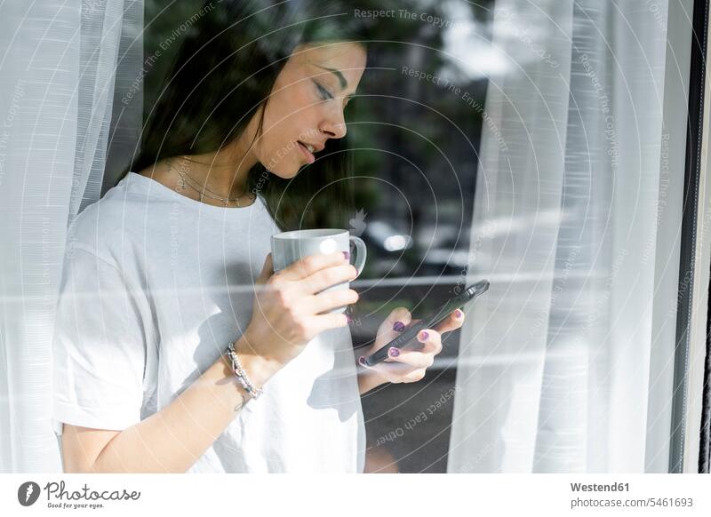Junge Frau mit Tasse Kaffee und Mobiltelefon hinter Fensterscheibe Leute Menschen People Person Personen Alleinstehende Alleinstehender Singles Unverheiratete