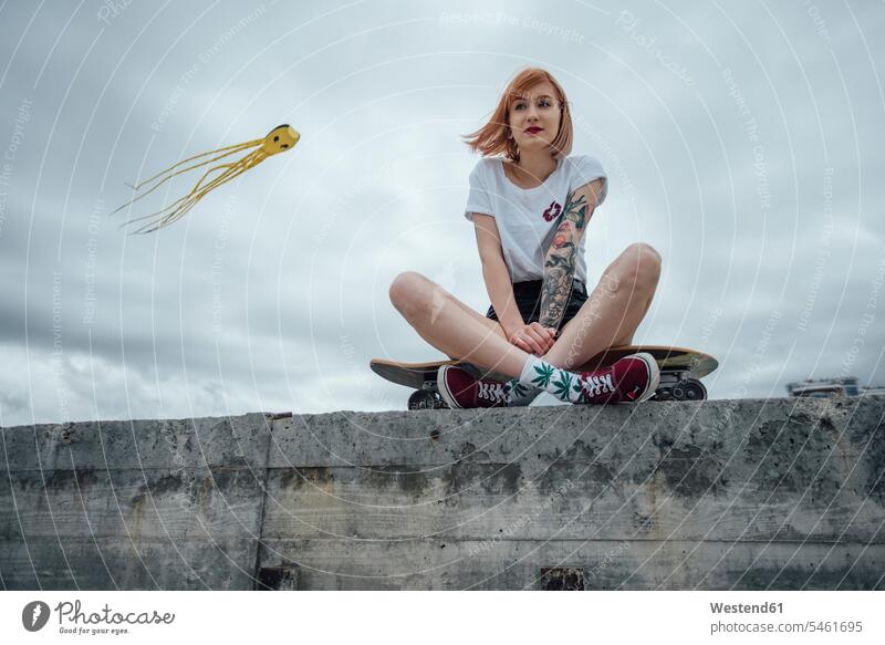 Junge Frau sitzt auf einer Betonwand auf Carver Skateboard mit Kite im Hintergrund Betonwände Betonwaende Drachen Rollbretter Skateboards weiblich Frauen sitzen