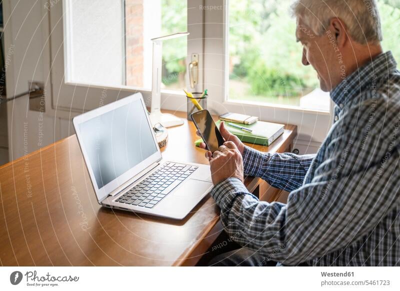 Älterer Mann sitzt zu Hause vor dem Laptop und benutzt ein Smartphone Leute Menschen People Person Personen Europäisch Kaukasier kaukasisch 1 Ein ein Mensch