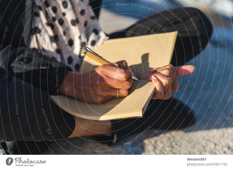 Frau sitzt an einer Wand und schreibt in ein Notizbuch, Nahaufnahme schreiben aufschreiben notieren schreibend Schrift Notizbücher Notizbuecher sitzen sitzend