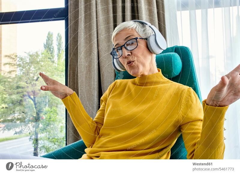 Ältere Frau sitzt im Sessel, hört Musik mit Kopfhörern Gardine Gardinen Vorhänge Stuehle Stühle Kopfhoerer Brillen hoeren sitzend freuen Frohsinn Fröhlichkeit